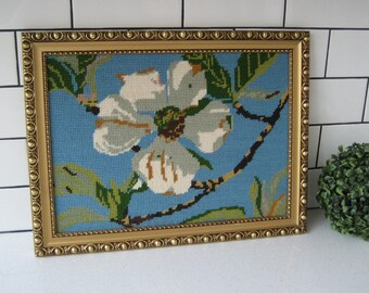 May include: A framed needlepoint featuring a white flower with brown and green details set against a light blue backdrop. The gold frame has a beaded border. The artwork is displayed on a white surface with a white tile background.