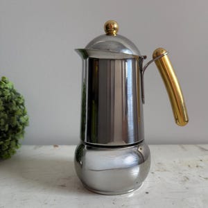 Op de afbeelding: Een zilveren en gouden espressomachine voor op het fornuis met een ronde basis en een gouden handvat.