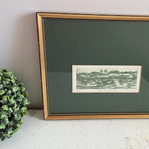 May include: A framed black and white print of a landscape scene with trees and a river. The frame is gold with a dark green mount.