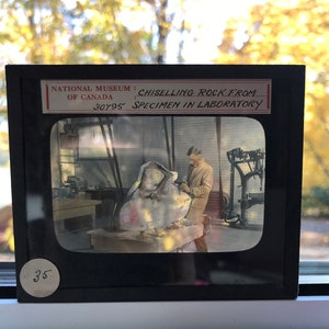 May include: A black and white photograph of a person chiseling a rock in a laboratory. The text on the image reads "NATIONAL MUSEUM: CHISELLING ROCK FROM... OF CANADA 30495 SPECIMEN IN LABORATORY".