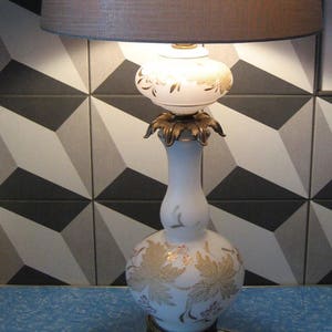 May include: A white ceramic table lamp with a gold leaf design and a brown fabric lampshade. The lamp has a brass base with four legs.