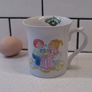 May include: White ceramic mug with a colorful illustration of two children sharing a sundae. The mug has the words "Cabbage Patch Kids" printed on it.