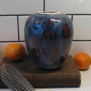 Könnte beinhalten: Eine blaue und orangefarbene Keramikvase mit abgerundeter Form steht auf einem alten Buch mit einer Feder daneben. Fünf Orangen liegen verstreut um die Vase und das Buch.