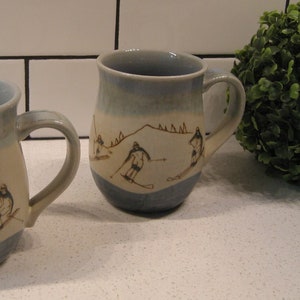 May include: Two ceramic mugs with a blue and white glaze. The mugs feature a brown illustration of skiers on a mountain.