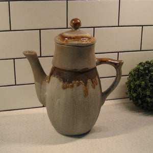 Peut inclure: Une théière en céramique brune et grise avec un couvercle et un glaçage brun goutte à goutte. La théière est posée sur un comptoir en carrelage blanc.