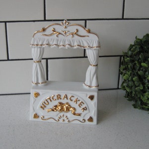 May include: White porcelain nutcracker figurine with a gold-painted design. The figurine depicts a small stage with curtains and the word "NUTCRACKER" written on the front.