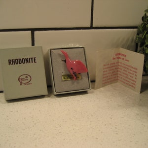 May include: A small pink bird figurine made of rhodonite, a stone known as the "Stone of Love." The figurine is displayed in a white box with a label that reads "Rhodonite" and "The Stone of Love." The box is sitting on a white tiled surface.