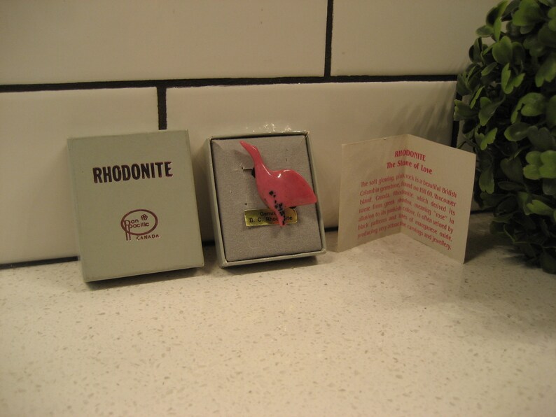May include: A small pink bird figurine made of rhodonite, a stone known as the "Stone of Love." The figurine is displayed in a white box with a label that reads "Rhodonite" and "The Stone of Love." The box is sitting on a white tiled surface.