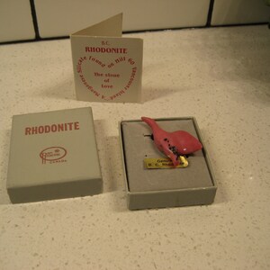 May include: A small pink rhodonite stone carved into the shape of a bird. The stone is in a small box with a label that reads "Genuine B.C. Rhodonite".