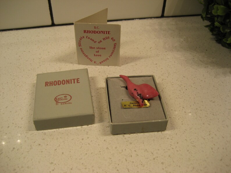 May include: A small pink rhodonite stone carved into the shape of a bird. The stone is in a small box with a label that reads "Genuine B.C. Rhodonite".