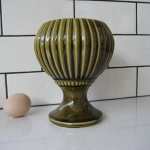 May include: A green ceramic planter with a fluted design and a pedestal base. The planter is on a white tiled surface.