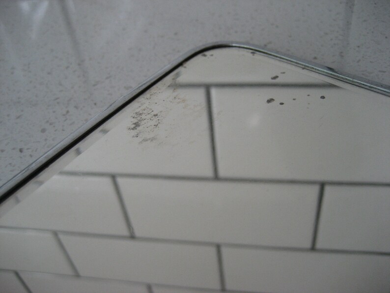 May include: A close-up of a silver-framed mirror with a few small water spots on the surface. The mirror is reflecting a white tiled wall.