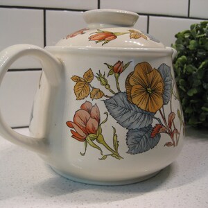 May include: A white ceramic teapot with a floral design. The teapot has a lid and a handle. The flowers are in shades of orange, yellow, and blue.