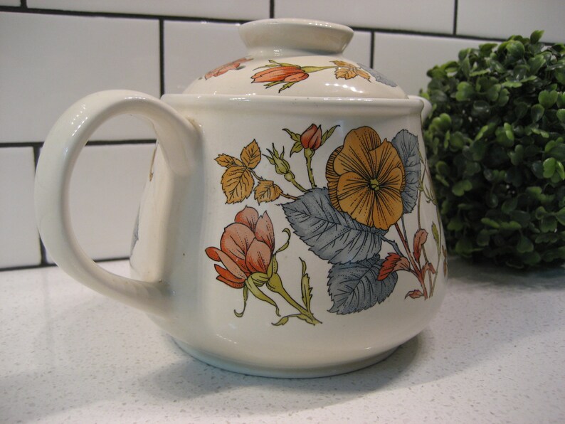 May include: A white ceramic teapot with a floral design. The teapot has a lid and a handle. The flowers are in shades of orange, yellow, and blue.