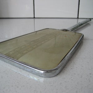 May include: A vintage hand mirror with a chrome frame and a pale yellow surface with a decorative pattern. The mirror has a long chrome handle.