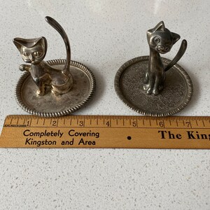 K&ouml;nnte beinhalten: Zwei silberne Katzen-Ringhalter. Jeder Halter zeigt eine Katzenfigur mit einem hochgestellten Schwanz, der als Ringhalter dient. Die Katzen sitzen auf kleinen, runden, silbernen Tabletts mit dekorativen Designs.