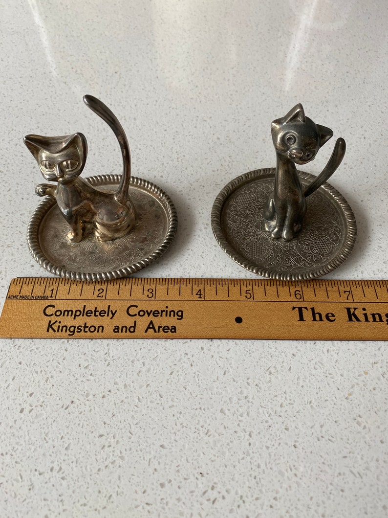 K&ouml;nnte beinhalten: Zwei silberne Katzen-Ringhalter. Jeder Halter zeigt eine Katzenfigur mit einem hochgestellten Schwanz, der als Ringhalter dient. Die Katzen sitzen auf kleinen, runden, silbernen Tabletts mit dekorativen Designs.