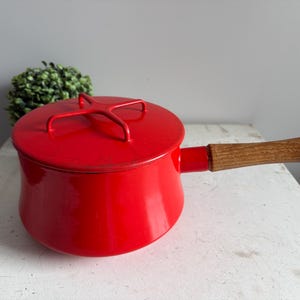 Puede incluir: Una cacerola de esmalte rojo con tapa y mango de madera. La tapa tiene un asa roja. La cacerola está sobre una superficie blanca. Una planta verde está en el fondo.