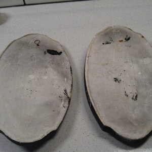 K&ouml;nnte beinhalten: Zwei ovale Wandplatten aus Keramik mit wei&szlig;em Hintergrund und schwarzen R&auml;ndern. Eine Platte hat einen kleinen Metallhaken oben. Beide Platten haben schwarze Farbflecken auf der Oberfl&auml;che.