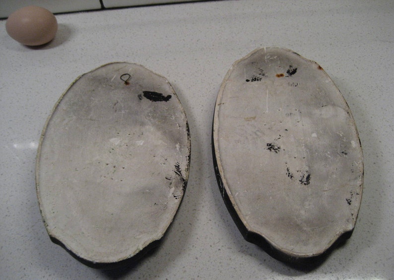 K&ouml;nnte beinhalten: Zwei ovale Wandplatten aus Keramik mit wei&szlig;em Hintergrund und schwarzen R&auml;ndern. Eine Platte hat einen kleinen Metallhaken oben. Beide Platten haben schwarze Farbflecken auf der Oberfl&auml;che.