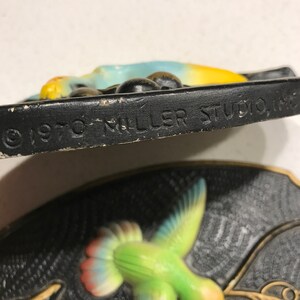 K&ouml;nnte beinhalten: Eine antike Wandplakette aus Keramik mit einem Kolibri im Flug mit gelbem Schnabel, gr&uuml;nem K&ouml;rper, blauen Fl&uuml;geln und roten Schwanzfedern. Die Plakette hat einen schwarzen Hintergrund mit einem goldfarbenen Zweig und dem Text "1970 Miller Studio Inc." unten.