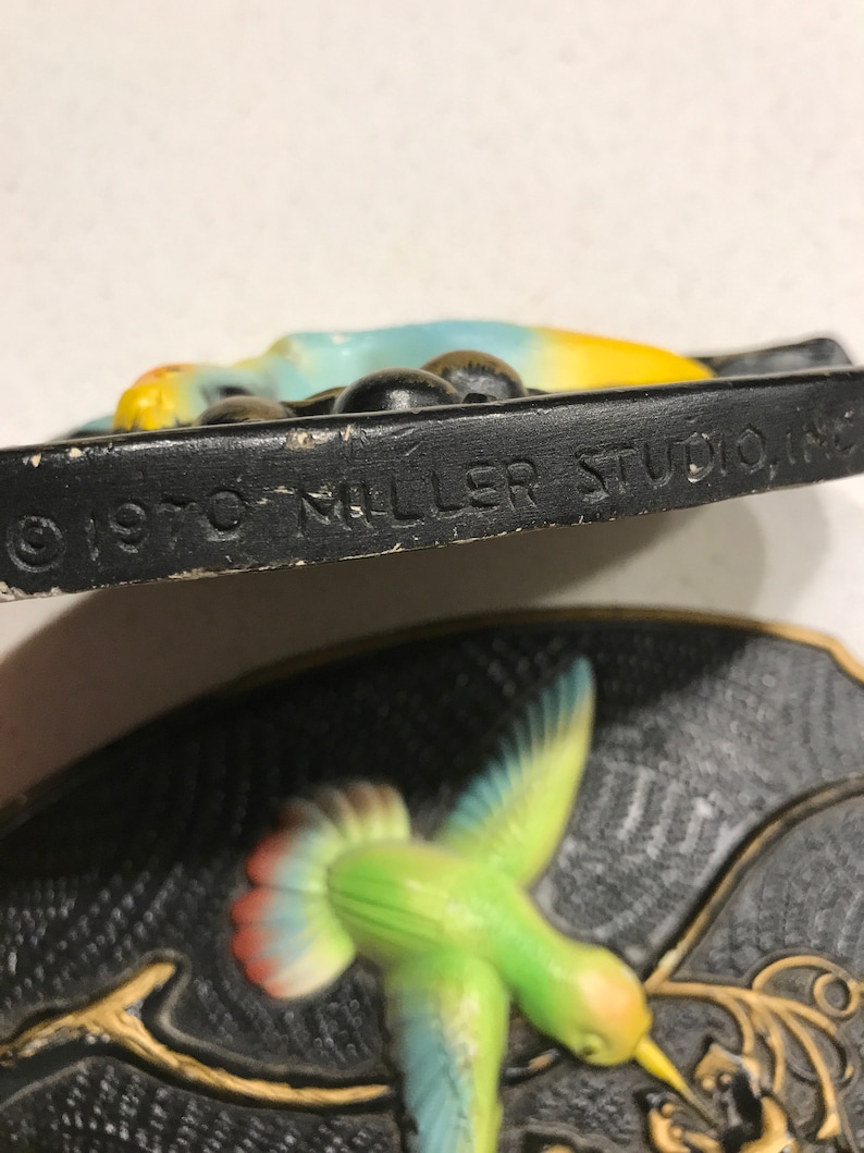 K&ouml;nnte beinhalten: Eine antike Wandplakette aus Keramik mit einem Kolibri im Flug mit gelbem Schnabel, gr&uuml;nem K&ouml;rper, blauen Fl&uuml;geln und roten Schwanzfedern. Die Plakette hat einen schwarzen Hintergrund mit einem goldfarbenen Zweig und dem Text "1970 Miller Studio Inc." unten.