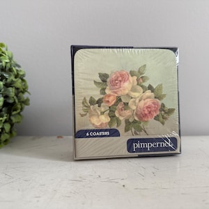 Op de afbeelding: Een doos met zes onderzetters met een bloemmotief van roze en crèmerozen en groene bladeren. De onderzetters zijn verpakt in een marineblauwe doos met de merknaam "pimpernel" erop.