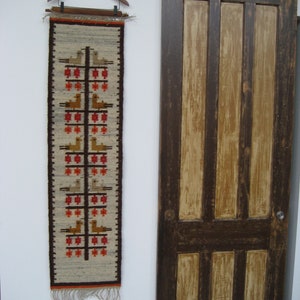 Puede incluir: Un tapiz de pared rectangular tejido con un patrón repetido de pájaros y formas geométricas en marrón, rojo y beige. El tapiz está suspendido de una percha de madera. A la derecha hay una puerta de madera marrón oscuro.