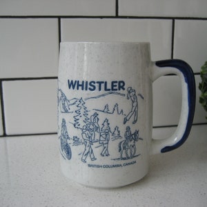 Puede incluir: Taza de cerámica blanca con borde y asa azules. La taza tiene una ilustración azul y blanca de personas esquiando y haciendo snowboard en Whistler, Columbia Británica, Canadá. El texto "WHISTLER" está impreso en azul en la taza.