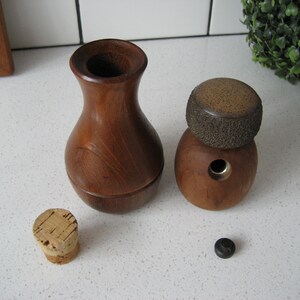 K&ouml;nnte beinhalten: Zwei Flaschenverschl&uuml;sse aus Holz, einer mit runder Oberseite und der andere mit einer vasenartigen Form. Beide Verschl&uuml;sse sind aus dunkelbraunem Holz mit nat&uuml;rlicher Maserung.