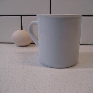 May include: A white ceramic mug with a handle on a white countertop. A white egg is in the background.