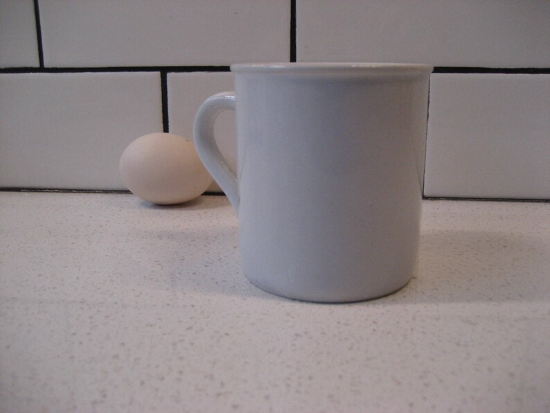 May include: A white ceramic mug with a handle on a white countertop. A white egg is in the background.