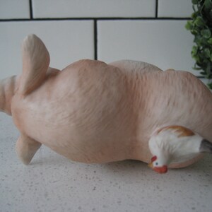 May include: A ceramic figurine of a light pink pig lying on its back with a small white and brown chicken peeking out from underneath. The pig has its front legs raised and its snout is visible. The figurine is on a white surface.