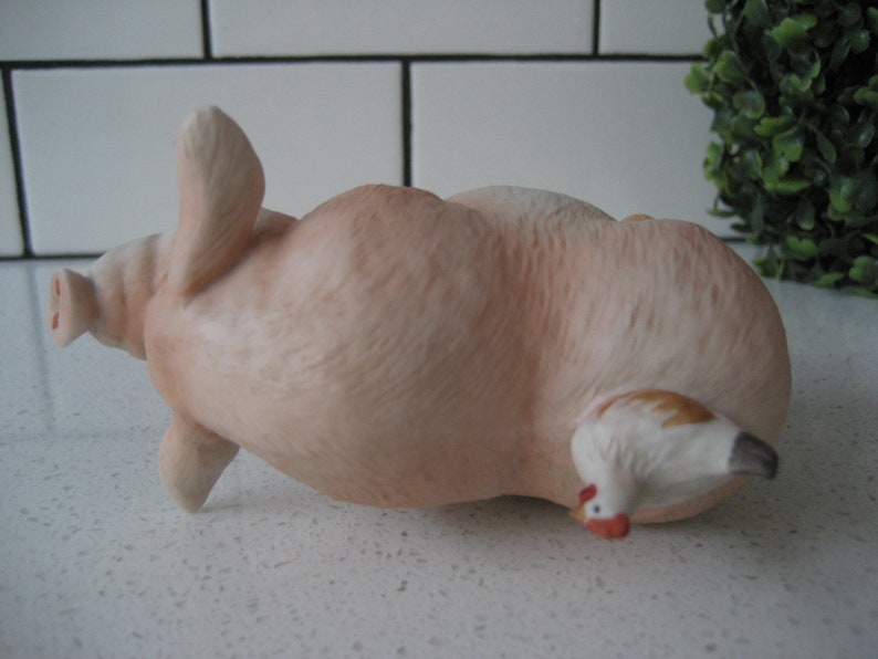 May include: A ceramic figurine of a light pink pig lying on its back with a small white and brown chicken peeking out from underneath. The pig has its front legs raised and its snout is visible. The figurine is on a white surface.