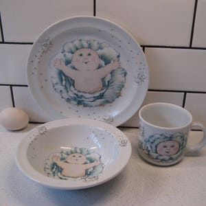 Peut inclure: Un ensemble de trois plats à thème Cabbage Patch Kids, comprenant une grande assiette, un bol plus petit et une tasse. Les plats sont blancs avec un motif de chou bleu et vert et présentent un bébé dessin animé avec un visage rose.