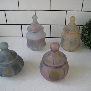 Puede incluir: Cuatro tarros de vidrio decorativos con tapas. Los tarros están esmerilados y tienen un efecto marmoleado en tonos azules, rosas y marrones. Los tarros están dispuestos sobre una superficie de azulejos blancos.