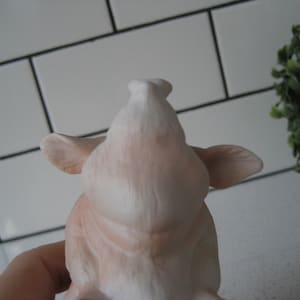 May include: A light pink ceramic pig figurine, viewed from the front, with visible ears and snout. The figurine is set against a white tiled background with a small green plant in the upper right corner.