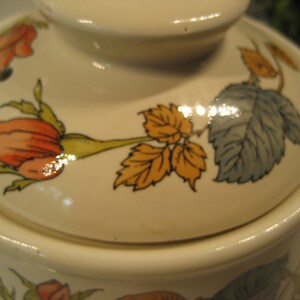 May include: A white ceramic teapot with a floral design featuring orange, yellow, and green roses and leaves. The lid has a glossy finish.