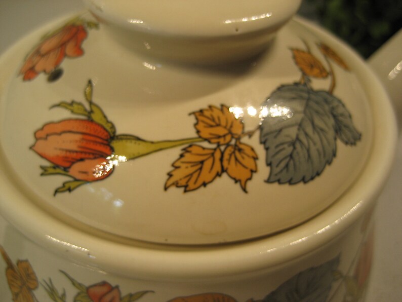 May include: A white ceramic teapot with a floral design featuring orange, yellow, and green roses and leaves. The lid has a glossy finish.
