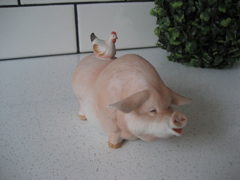 May include: A ceramic figurine of a smiling pink pig with a small chicken perched on its back. The pig has a detailed snout and small ears. The chicken is white and brown with a red comb. The figurine is a decorative item.