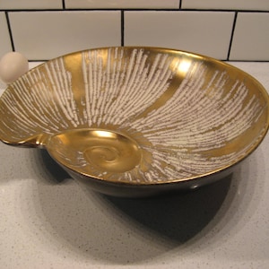 Puede incluir: Un cuenco de cerámica dorado y blanco con forma de concha. El cuenco tiene una superficie texturizada y un diseño en relieve.