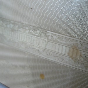 May include: Close-up of a silver-colored metal surface with an intricate engraved design. The design features a series of wavy lines, a geometric pattern, and a floral motif.