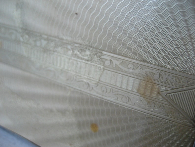 May include: Close-up of a silver-colored metal surface with an intricate engraved design. The design features a series of wavy lines, a geometric pattern, and a floral motif.