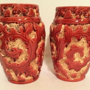 May include: Two red ceramic vases with intricate white floral designs. The vases have a textured surface and a rounded shape.