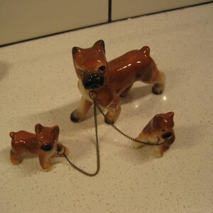 May include: Three ceramic figurines of brown and white boxer dogs. The largest dog is standing and has a chain attached to its collar. The two smaller dogs are sitting and are connected to the chain.
