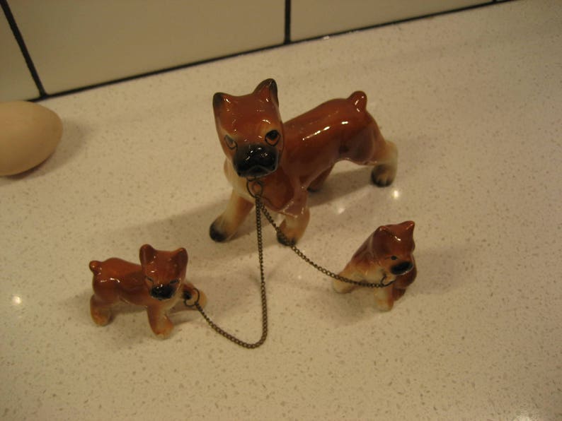May include: Three ceramic figurines of brown and white boxer dogs. The largest dog is standing and has a chain attached to its collar. The two smaller dogs are sitting and are connected to the chain.
