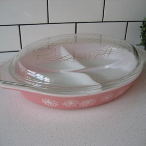 Op de afbeelding: Roze ovale ovenschaal met een doorzichtig glazen deksel. De schaal heeft een wit bloemmotief rond de rand en een gouden swirl-ontwerp op het deksel.