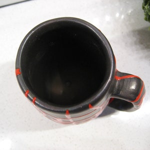 K&ouml;nnte beinhalten: Eine schwarze Keramiktasse mit rotem Streifenmuster. Die Tasse ist leer und hat ein dunkles Inneres.