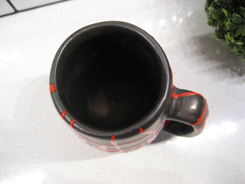 K&ouml;nnte beinhalten: Eine schwarze Keramiktasse mit rotem Streifenmuster. Die Tasse ist leer und hat ein dunkles Inneres.
