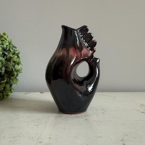 Könnte beinhalten: Eine dunkelbraune und violette Keramikvase mit einem einzigartigen handförmigen Design. Die Vase hat eine runde Öffnung oben und eine glatte, glänzende Oberfläche.